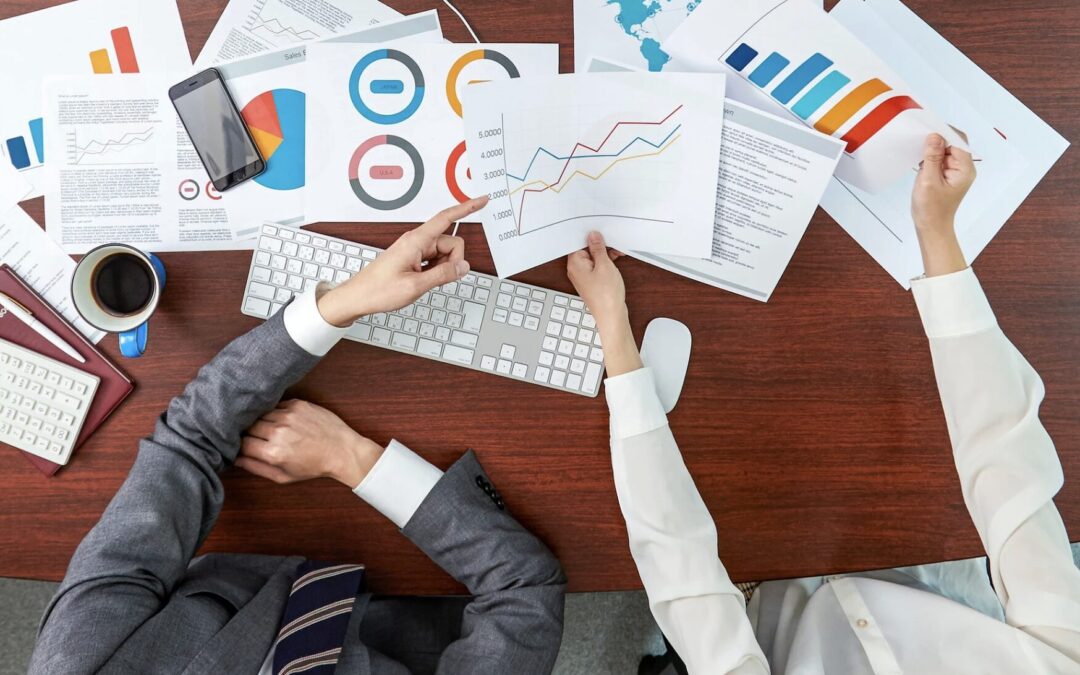 A man and a woman analyze sheets of paper with printed graphs and charts sitting on a desk.